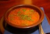 Tomato soup with green herbs in a brown bowl sitting on a blue cloth on a dining table. 
