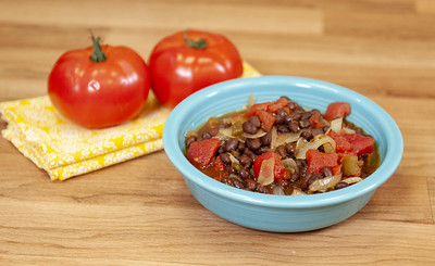 Black bean and tomato soup with onions inside a blue bowl. Two whole tomatoes sitting on a clothe napkin behind the bowl of soup. 