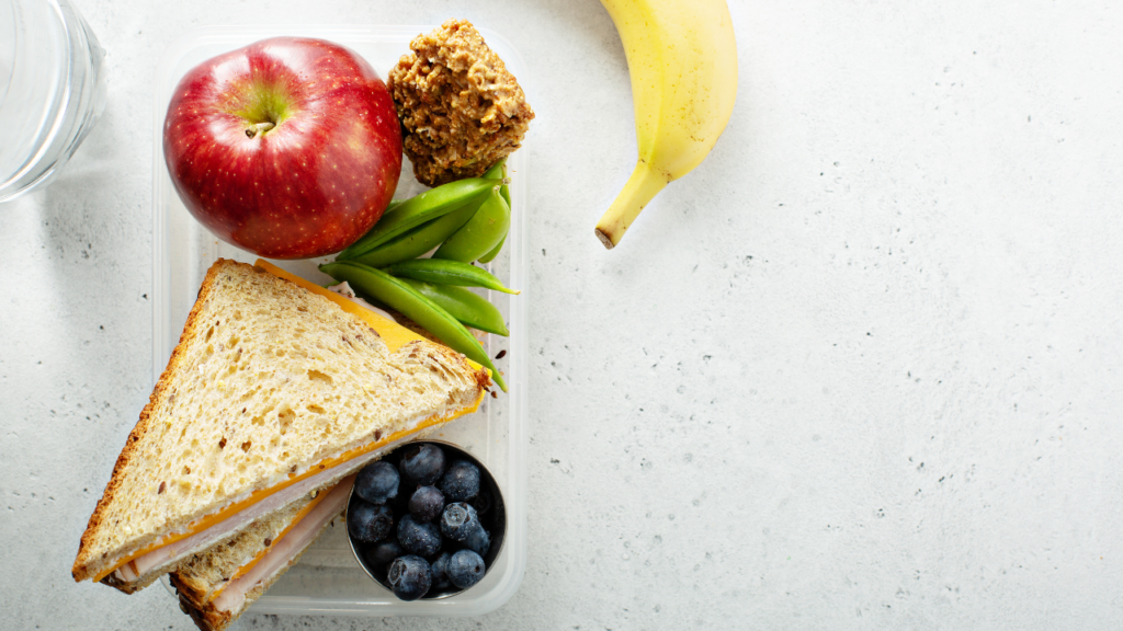 A healthy lunchbox containing a turkey sandwich, granola, peas and various fruits.