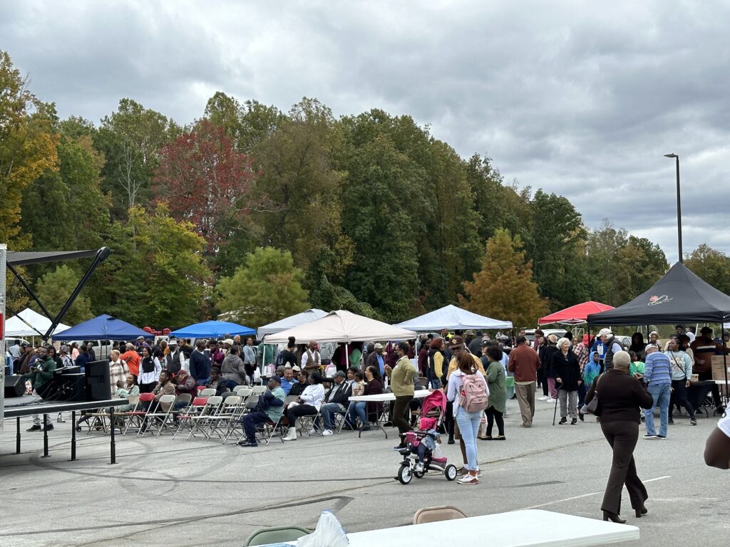 A mass of attendees are pictured at a distance, walking between tents at the farmer's market. 