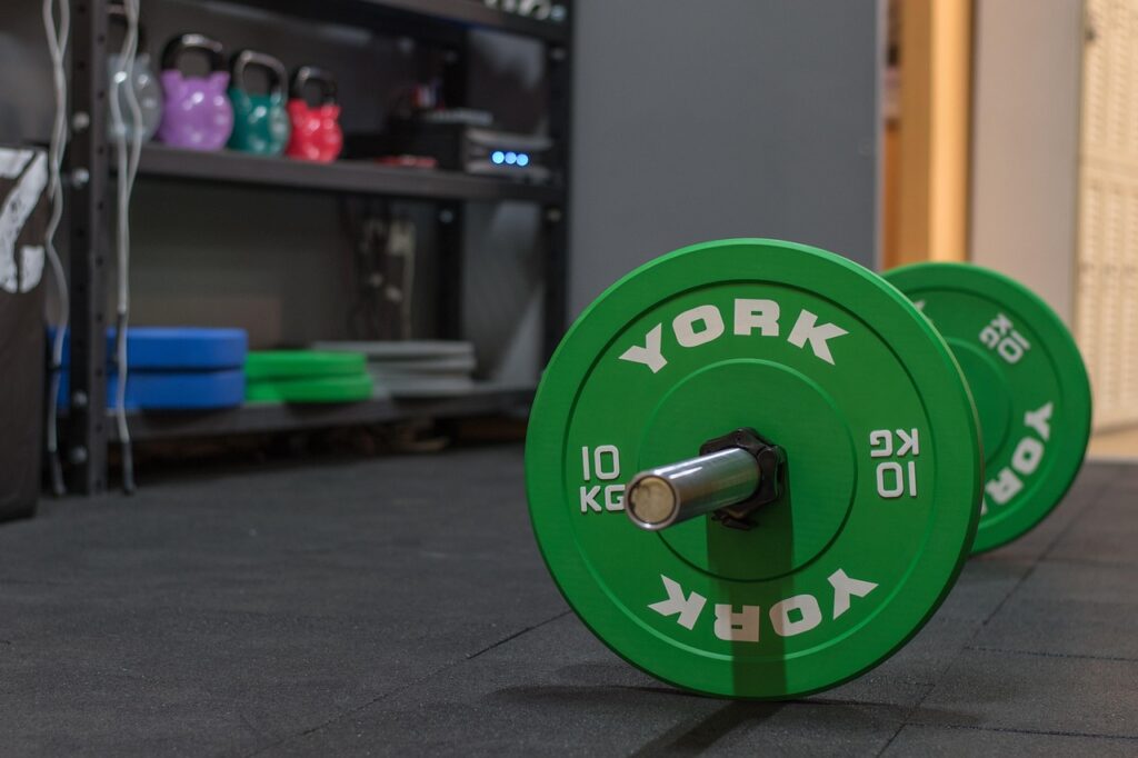 Barbell with weight on it and shelf with weight plates and kettlebells. 